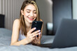 © F8  \ Suport Ukraine - Beautiful smiling woman lying on the bed by the laptop and typing a message on her phone.