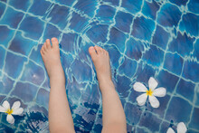Above Shot With Copy Space Of Asian Girl's Legs Sitting Beside The Pool With Blue, Clear Water And Oriental Flowers Under Sunshine With Fun And Happiness During The Summer Vacation With The Family.