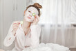 © Sviatlana - A cute girl with a face mask covers one eye with a slice of cucumber, holds the second cucumber in her hand and sits on the bed in the bedroom. The concept of beauty and health
