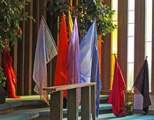 This Is Several Sheer And Shimmery Colored, Plain Flags Sitting Next To Some Stained Glass Windows Indoors With Sunlight Hitting Them.