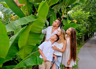 Cheerful, happy family, father, mother, son and daughter enjoying walk in banana palm leaves. Family in white clothes having fun on walkway with big palms tree. Summer day and vacation time concept.