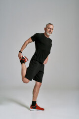 Full length shot of middle aged muscular man in black sportswear looking at camera while stretching his body before workout in studio over grey background