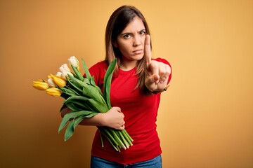 Poster - Young blonde woman holding romantic bouquet of tulips flowers over yellow background Pointing with finger up and angry expression, showing no gesture