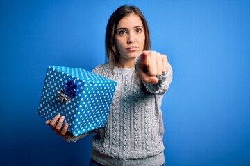 Sticker - Young blonde woman holding a gift box as birthday present over isolated background pointing with finger to the camera and to you, hand sign, positive and confident gesture from the front