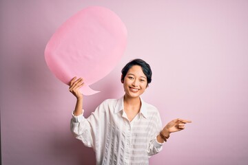 Poster - Young beautiful asian girl talking holding speech bubble over isolated pink background very happy pointing with hand and finger to the side