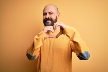 Sticker - Handsome bald man with beard wearing casual sweater standing over yellow background smiling in love doing heart symbol shape with hands. Romantic concept.