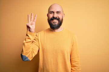 Canvas Print - Handsome bald man with beard wearing casual sweater standing over yellow background showing and pointing up with fingers number three while smiling confident and happy.