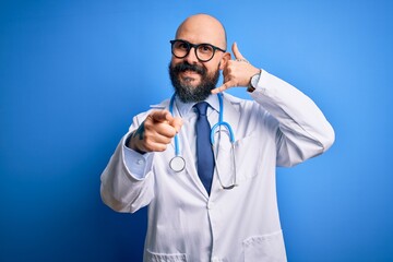 Sticker - Handsome bald doctor man with beard wearing glasses and stethoscope over blue background smiling doing talking on the telephone gesture and pointing to you. Call me.