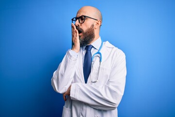 Wall Mural - Handsome bald doctor man with beard wearing glasses and stethoscope over blue background bored yawning tired covering mouth with hand. Restless and sleepiness.