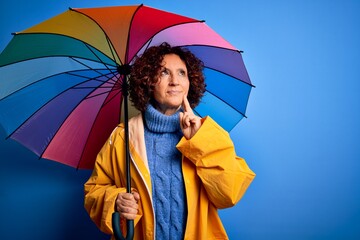 Poster - Middle age curly hair woman wearing rain coat holding colorful umbrella over blue background serious face thinking about question, very confused idea