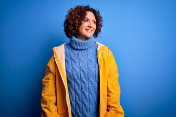 Poster - Middle age curly hair woman wearing rain coat standing over isolated blue background looking away to side with smile on face, natural expression. Laughing confident.