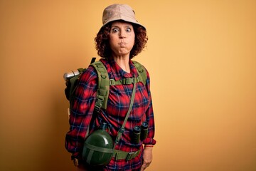 Wall Mural - Middle age curly hair hiker woman hiking wearing backpack and water canteen using binoculars puffing cheeks with funny face. Mouth inflated with air, crazy expression.