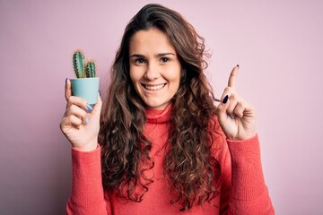 Sticker - Young beautiful woman with curly hair holding small cactus over isolated pink background surprised with an idea or question pointing finger with happy face, number one