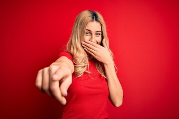 Poster - Young beautiful blonde woman wearing casual t-shirt standing over isolated red background laughing at you, pointing finger to the camera with hand over mouth, shame expression