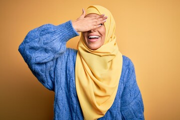 Canvas Print - Young beautiful brunette muslim woman wearing arab hijab over isolated yellow background smiling and laughing with hand on face covering eyes for surprise. Blind concept.