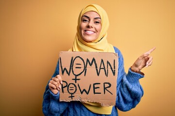 Wall Mural - Young beautiful arab woman wearing islamic hijab holding banner with women power message very happy pointing with hand and finger to the side