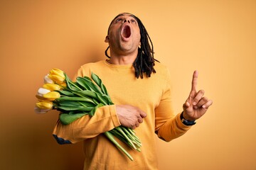 Wall Mural - Young african american afro romantic man with dreadlocks holding bouquet of yellow tulips amazed and surprised looking up and pointing with fingers and raised arms.