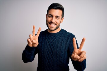 Young handsome man with beard wearing casual sweater standing over white background smiling looking to the camera showing fingers doing victory sign. Number two.