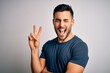 Young handsome man wearing casual t-shirt standing over isolated white background smiling with happy face winking at the camera doing victory sign. Number two.