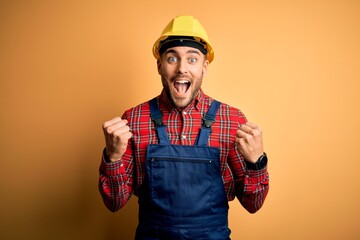 Young builder man wearing construction uniform and safety helmet over yellow isolated background celebrating surprised and amazed for success with arms raised and open eyes. Winner concept.