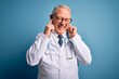 © Krakenimages.com - Senior grey haired doctor man wearing stethoscope and medical coat over blue background covering ears with fingers with annoyed expression for the noise of loud music. Deaf concept.