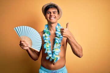 Poster - Young tourist man on vacation wearing swimwear and hawaiian lei flowers holding hand fan happy with big smile doing ok sign, thumb up with fingers, excellent sign