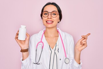 Wall Mural - Young beautiful doctor woman with blue eyes wearing stethoscope holding medicine pills very happy pointing with hand and finger to the side