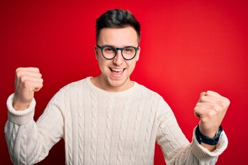 Wall Mural - Young handsome caucasian man wearing glasses and casual winter sweater over red background celebrating surprised and amazed for success with arms raised and open eyes. Winner concept.