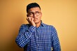 © Krakenimages.com - Young handsome latin man wearing casual shirt and glasses over yellow background looking stressed and nervous with hands on mouth biting nails. Anxiety problem.