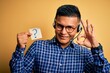 © Krakenimages.com - Young latin call center agent man holding question mark reminder paper using headset doing ok sign with fingers, excellent symbol