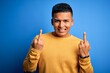 © Krakenimages.com - Young handsome latin man wearing yellow casual sweater over isolated blue background Showing middle finger doing fuck you bad expression, provocation and rude attitude. Screaming excited