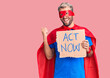 Young blond man wearing super hero costume holding act now cardboard banner pointing thumb up to the side smiling happy with open mouth