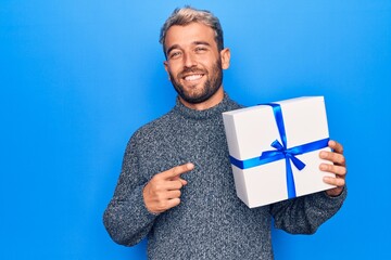 Young handsome blond man with beard holding birthday present over isolated blue background smiling happy pointing with hand and finger