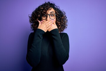 Poster - Young beautiful curly arab woman wearing casual sweater and glasses over purple background shocked covering mouth with hands for mistake. Secret concept.