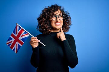 Poster - Young beautiful curly arab woman wearing glasses holding uk flag over blue background serious face thinking about question, very confused idea