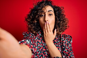 Poster - Young beautiful curly arab woman wearing dress making selfie by camera over red background cover mouth with hand shocked with shame for mistake, expression of fear, scared in silence, secret concept