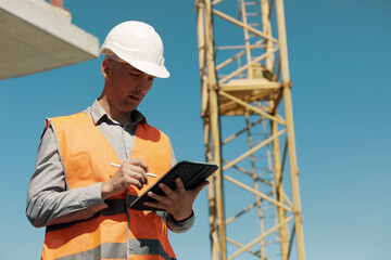 An engineer in an orange vest and a white construction control helmet conducts an inspection with a tablet in his hands against the background of a construction site and a tower crane