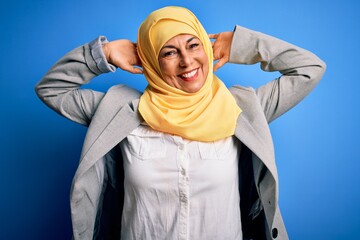 Sticker - Middle age brunette business woman wearing muslim traditional hijab over blue background relaxing and stretching, arms and hands behind head and neck smiling happy