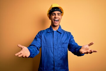 Wall Mural - Young handsome african american worker man wearing blue uniform and security helmet smiling cheerful offering hands giving assistance and acceptance.