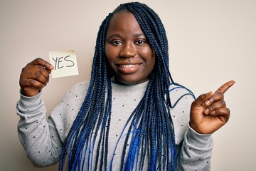 Sticker - Young african american plus size woman with braids holding reminder paper with yes message very happy pointing with hand and finger to the side