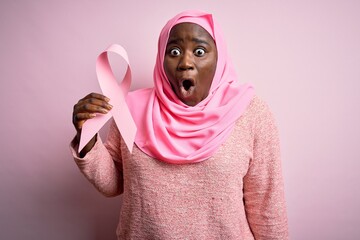 Poster - Young african american plus size woman wearing muslim hijab holding pink cancer ribbon scared in shock with a surprise face, afraid and excited with fear expression