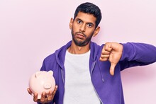 Young Latin Man Holding Piggy Bank With Angry Face, Negative Sign Showing Dislike With Thumbs Down, Rejection Concept