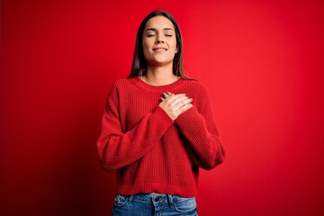 Wall Mural - Beautiful young brunette woman wearing casual sweater standing over red isolated background smiling with hands on chest with closed eyes and grateful gesture on face. Health concept.