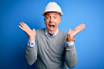 Poster - Middle age handsome grey-haired engineer man wearing safety helmet over blue background celebrating crazy and amazed for success with arms raised and open eyes screaming excited. Winner concept