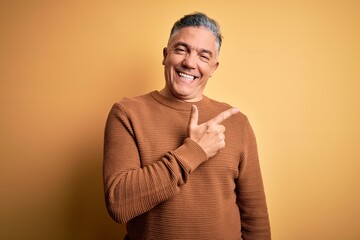 Wall Mural - Middle age handsome grey-haired man wearing casual sweater over yellow background cheerful with a smile of face pointing with hand and finger up to the side with happy and natural expression on face