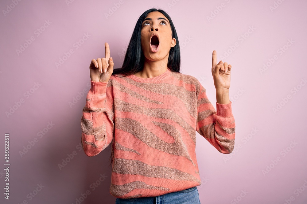 Young beautiful hispanic fashion woman wearing animal print sweater over pink background amazed and surprised looking up and pointing with fingers and raised arms.