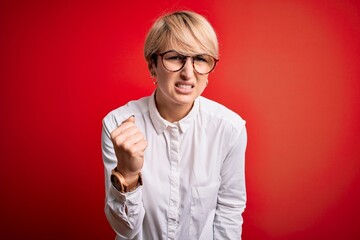 Canvas Print - Young blonde business woman with short hair wearing glasses over red background angry and mad raising fist frustrated and furious while shouting with anger. Rage and aggressive concept.