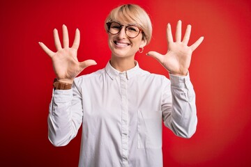 Canvas Print - Young blonde business woman with short hair wearing glasses over red background showing and pointing up with fingers number ten while smiling confident and happy.