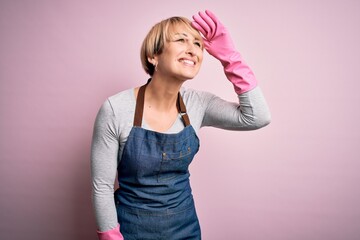 Poster - Young blonde cleaner woman with short hair wearing apron and gloves over pink background very happy and smiling looking far away with hand over head. Searching concept.