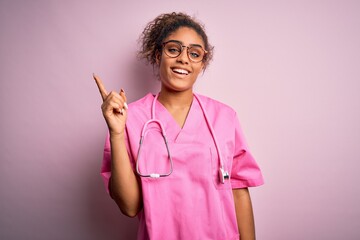 Poster - African american nurse girl wearing medical uniform and stethoscope over pink background with a big smile on face, pointing with hand and finger to the side looking at the camera.